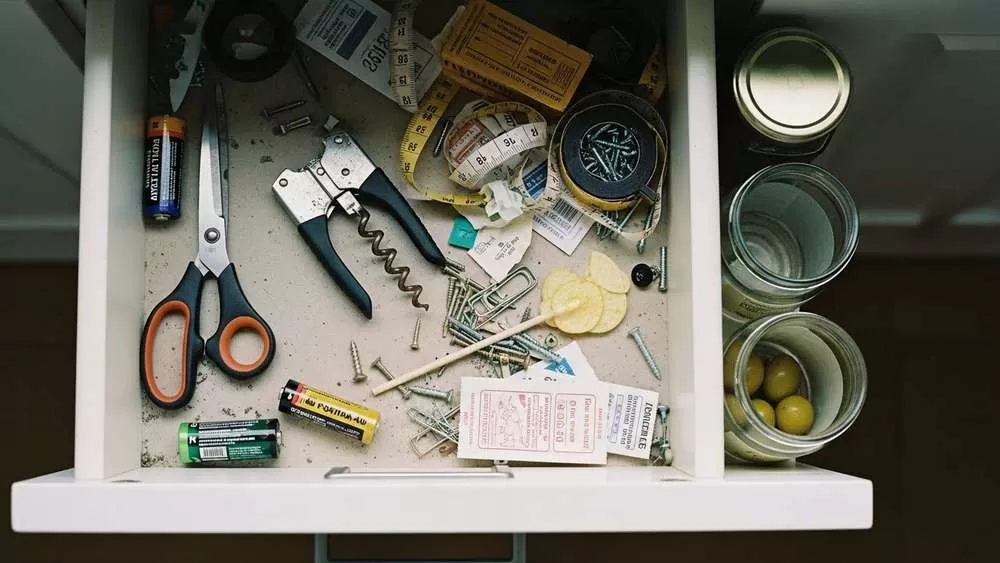 Vista superior da gaveta bagunçada da cozinha com itens inúteis e poeira, ao lado de potes de vidro vazios e tesoura para solução de organização DIY sem custo.